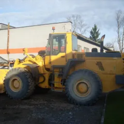 Żółta maszyna budowlana z dużą łyżką z przodu, stojąca na ziemi obok budynku z pomarańczowym pasem. Widoczne drzewa w tle.