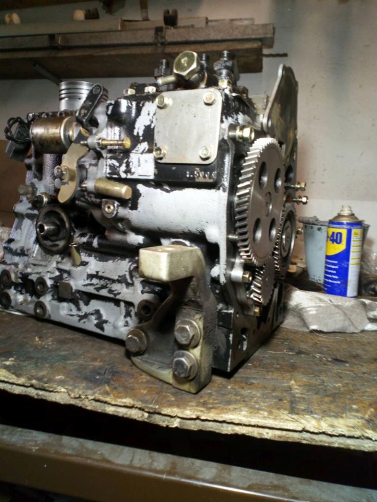 Częściowo rozmontowany silnik maszyny budowlanej na warsztatowym stole, widoczne koła zębate, fragment tłoka i puszka WD-40 w tle.