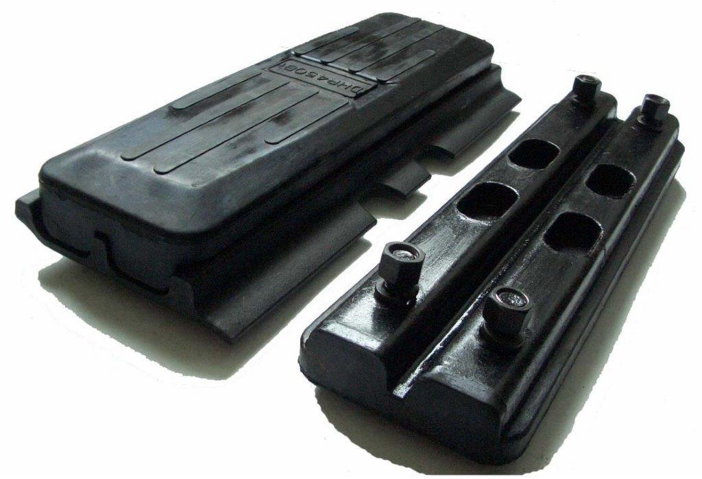 Dwa czarne, gumowe elementy maszyny budowlanej, prawdopodobnie podkładki lub odbojniki, leżące na białym tle. Jeden element jest prostokątny z podłużnymi rowkami, drugi składa się z dwóch...