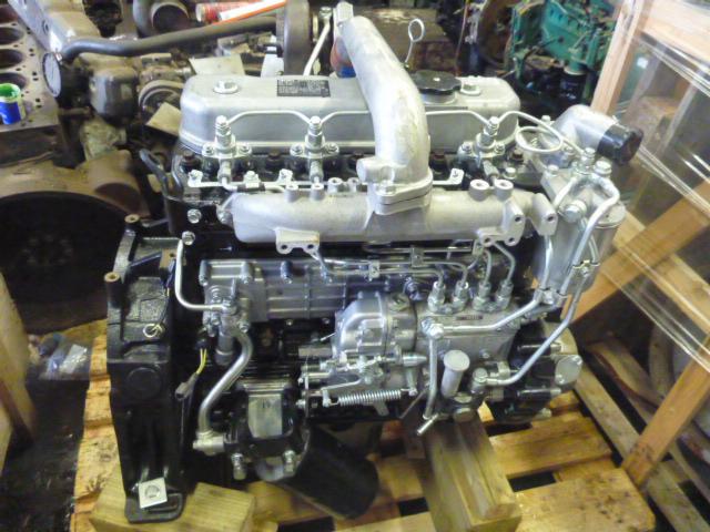 Szczegółowe ujęcie silnika maszyny budowlanej, stojącego na drewnianych podporach, z widocznymi licznymi przewodami i metalowymi elementami, na tle innych maszyn i elementów warsztatowych.