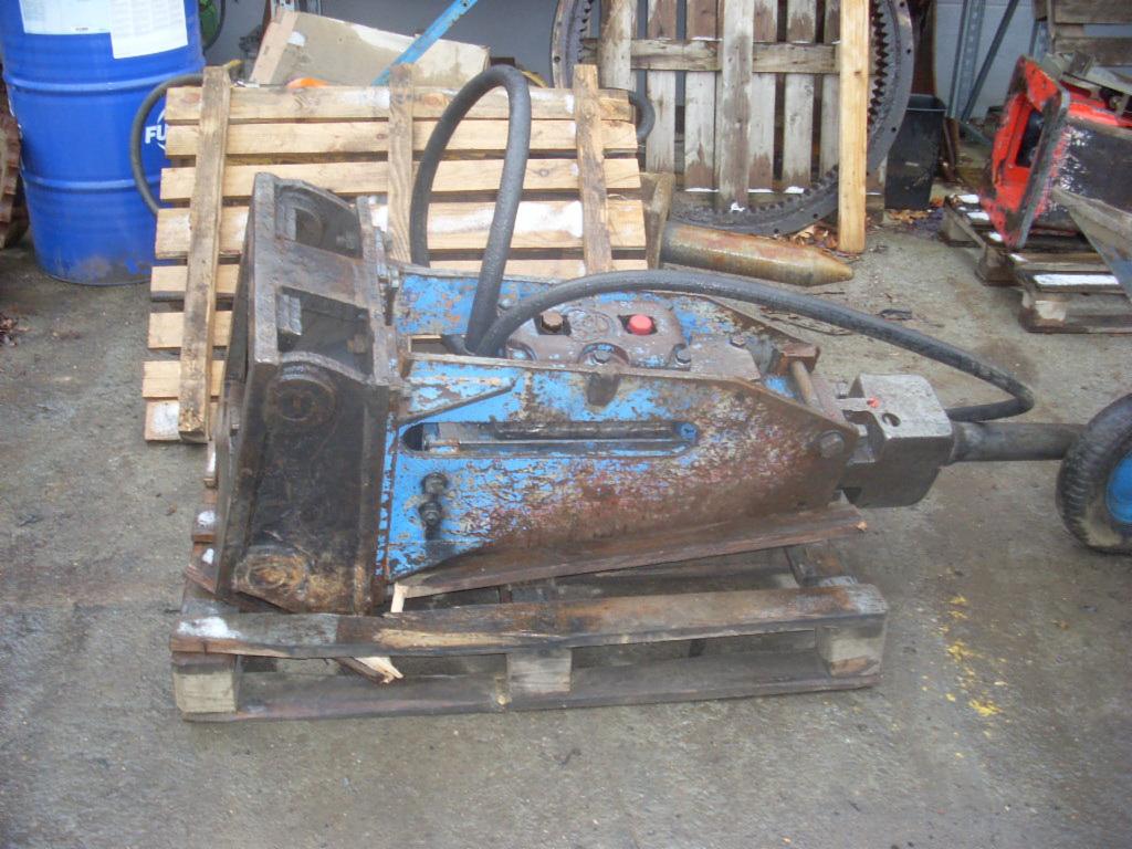 Hydrauliczny młot wyburzeniowy, pomalowany na niebiesko, z widocznymi śladami korozji i użytkowania, umieszczony na drewnianej palecie. W tle widoczna niebieska beczka i drewniane elementy.