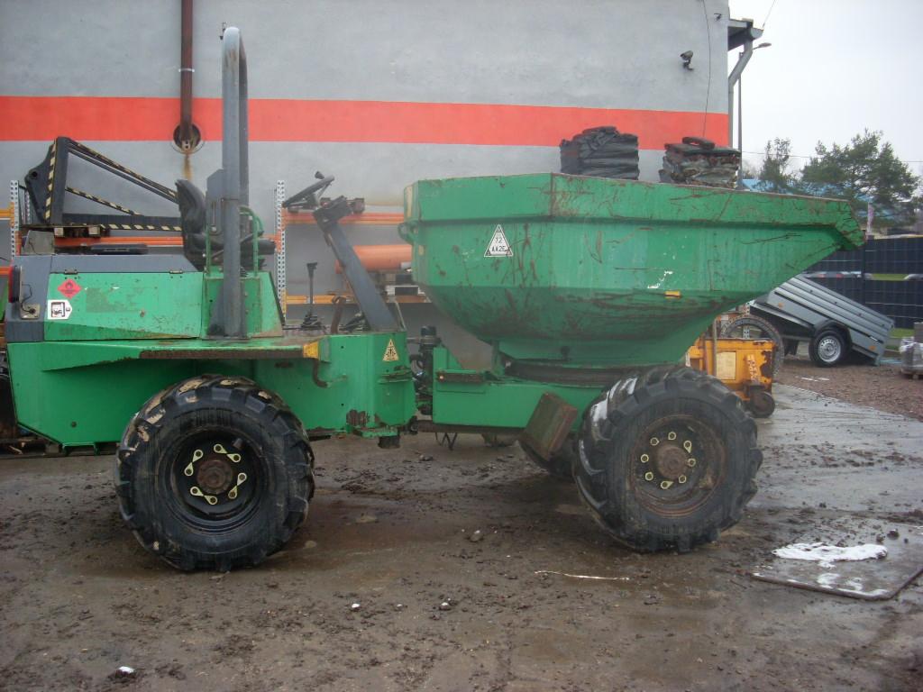 Zielony wywrotka budowlana z widocznymi śladami użytkowania, stojąca na błotnistym terenie, z elementami rusztowania i przyczepą w tle.