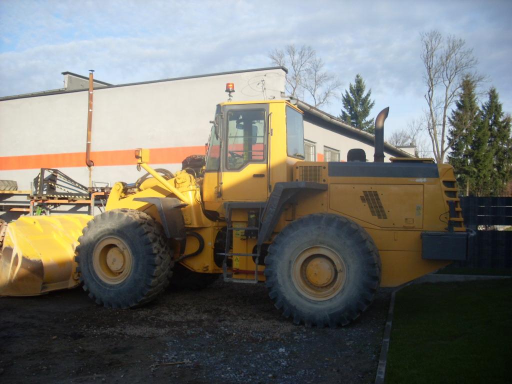 Żółta maszyna budowlana z dużą łyżką z przodu, stojąca na ziemi obok budynku z pomarańczowym pasem. Widoczne drzewa w tle.