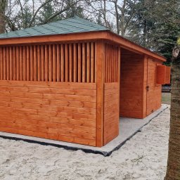 Midgard - Nowoczesna, drewniana konstrukcja z zielonym dachem, prawdopodobnie domek ogrodowy lub pawilon, wzniesiona na betonowej podstawie wysypanej piaskiem, z widocznymi pionowymi szczeblami i zamkniętymi...