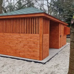 Nowoczesna, drewniana konstrukcja z zielonym dachem, prawdopodobnie domek ogrodowy lub pawilon, wzniesiona na betonowej podstawie wysypanej piaskiem, z widocznymi pionowymi szczeblami i zamkniętymi...