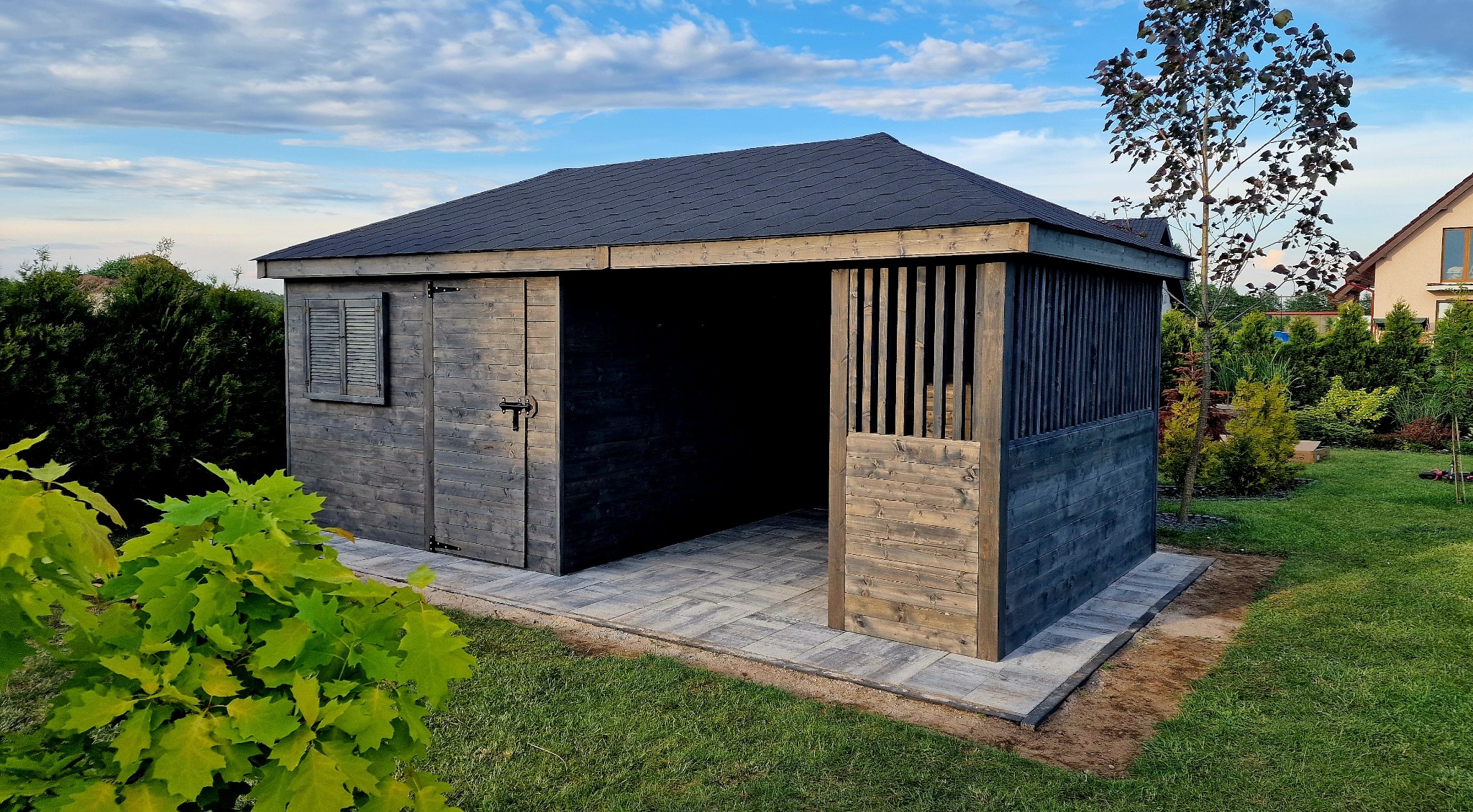 Drewniany domek ogrodowy o powierzchni 18m2 z przesuwnymi drzwiami i okiennicą, wykończony w szarym odcieniu, na tle zielonego trawnika i błękitnego nieba.
