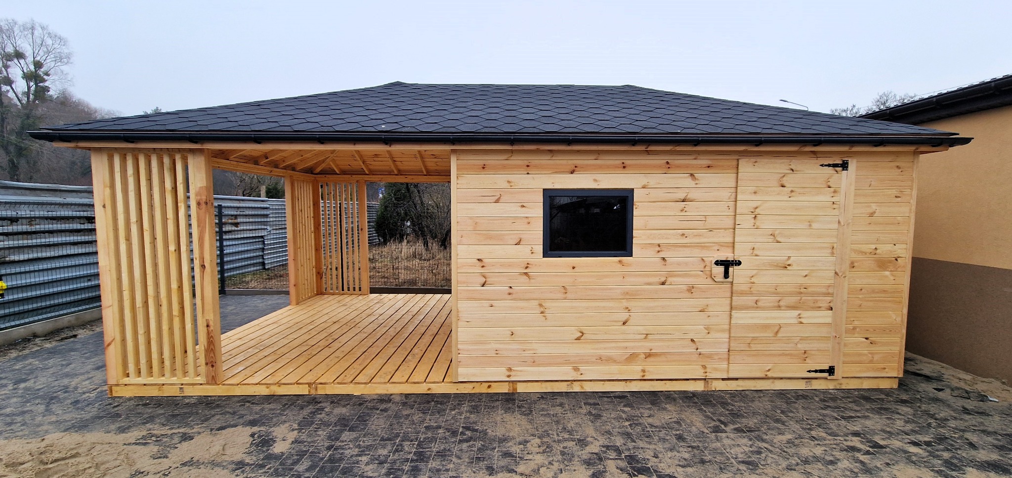 Domek ogrodowy z jasnego drewna z zadaszoną częścią tarasową i zamkniętą częścią z oknem i drzwiami, kryty ciemną dachówką bitumiczną.