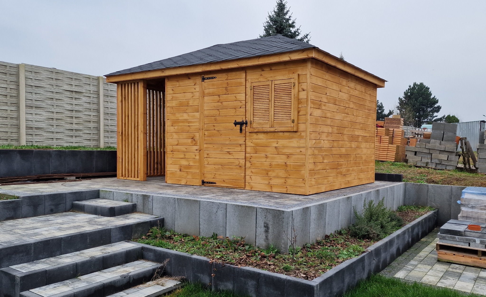 Nowoczesny, drewniany domek ogrodowy z czarnym dachem, zamykanymi okiennicami i ażurową ścianą, posadowiony na podwyższeniu z szarej kostki brukowej, z widocznymi schodami i elementami...