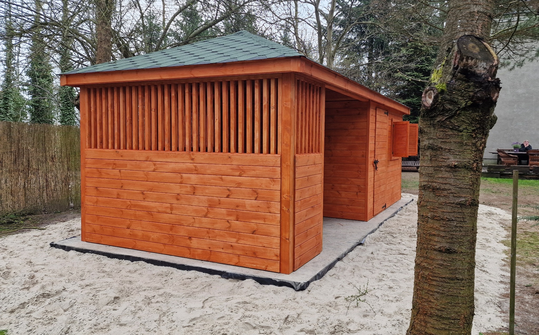 Nowoczesna, drewniana konstrukcja z zielonym dachem, prawdopodobnie domek ogrodowy lub pawilon, wzniesiona na betonowej podstawie wysypanej piaskiem, z widocznymi pionowymi szczeblami i zamkniętymi...