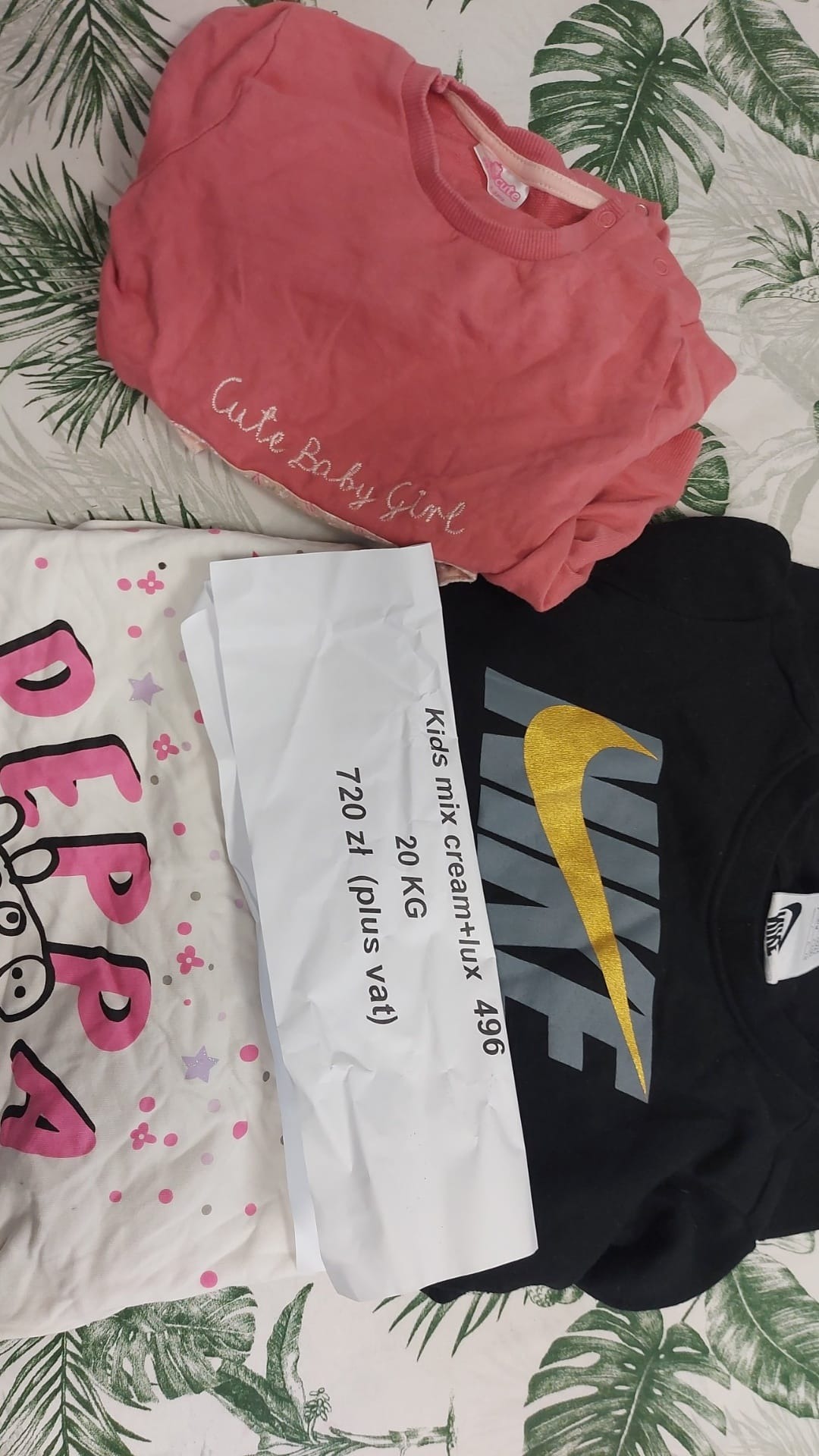 Stos ubrań dziecięcych, w tym różowa bluza z napisem 'Cute Baby Girl', czarna bluza Nike z logo, oraz biała koszulka z motywem Peppy Pig, obok faktura za 20kg odzieży 'Kids mix cream+lux 496' za...
