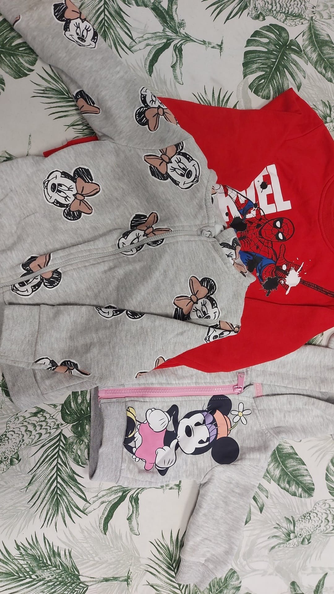 Rozłożone na wzorzystej pościeli szare bluzy z motywami Myszki Minnie i czerwona koszulka z nadrukiem Spider-Mana.