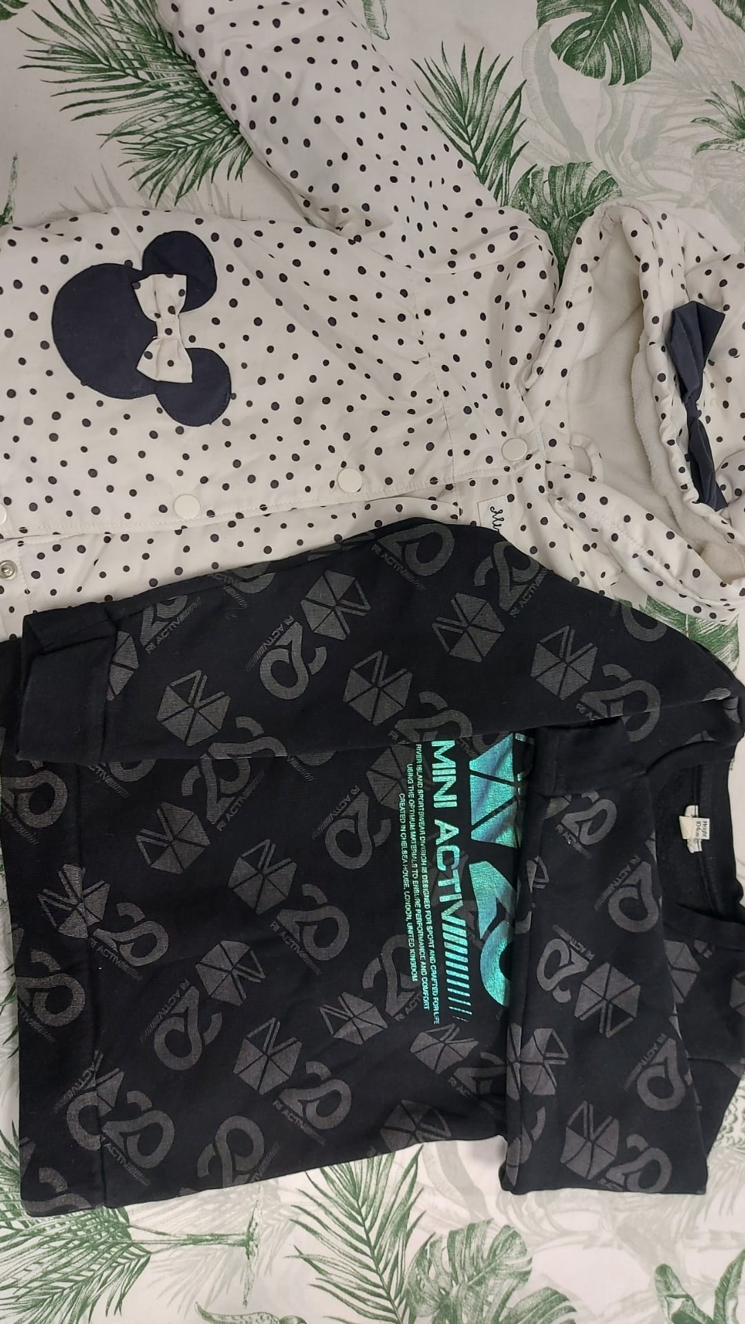 Dwie dziecięce kurtki, jedna biała w czarne kropki z aplikacją Myszki Miki, druga czarna z nadrukiem 'MINI ACTIVE' i wzorami geometrycznymi, ułożone na pościeli w tropikalne liście.