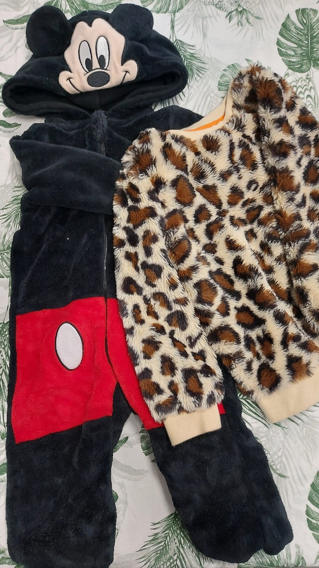 Czarny pluszowy kombinezon z kapturem Myszki Miki obok swetra w panterkę na tle w liście.