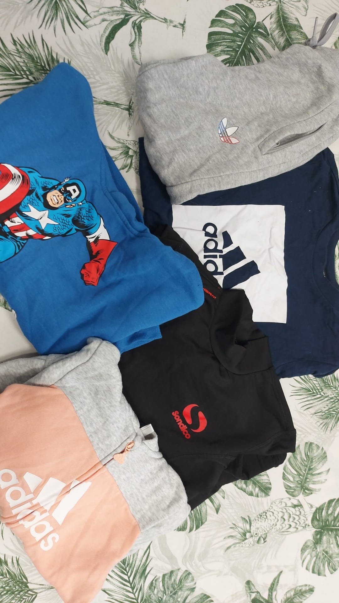 Stos pogniecionych ubrań różnych marek i wzorów leżący na wzorzystym tle, w tym niebieska bluza z Kapitanem Ameryką, szara bluza Adidas, granatowa koszulka z logo Adidas i czarna bluza Sonadico.