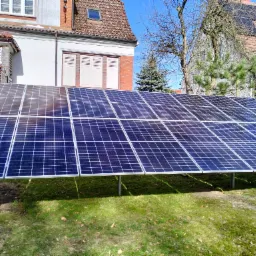 Zestaw paneli słonecznych ustawiony na trawniku przed domem z czerwoną dachówką i oknami z białymi okiennicami, widoczne drzewa bez liści i iglak.