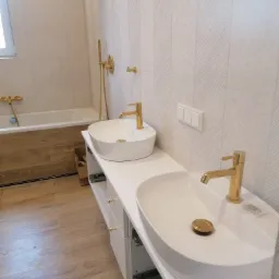 Nowoczesna łazienka z dwoma białymi umywalkami nablatowymi o owalnym kształcie, złotą armaturą i drewnopodobną obudową wanny, wykończona płytkami w odcieniach bieli z delikatnym wzorem jodełki.