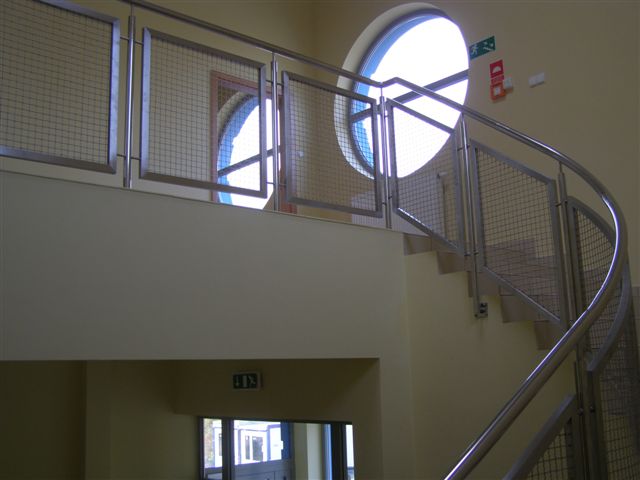 Spiralne schody z balustradą ze stali nierdzewnej z siatkowym wypełnieniem, okrągłe okno w tle, widok z dołu.