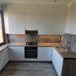 Biała kuchnia na wymiar z drewnianym blatem, widok na blok mieszkalny przez okno, szare płytki nad blatem, płyta gazowa i piekarnik.