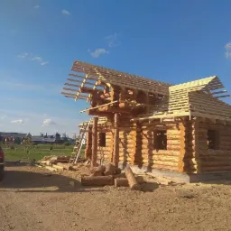 Budowa domu z bali drewnianych, widoczna konstrukcja dachu z łatami, drabina oparta o ścianę, osoba przy samochodzie w tle, teren budowy.