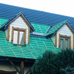 Pracownik montuje ciemne panele na dachu domu z dwoma lukarnami, widoczne zielone membrany i drewniana konstrukcja dachu, częściowo pokryty dach, narzędzia i wiadro na dachu.