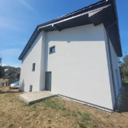 Nowoczesny, dwukondygnacyjny dom z białymi ścianami elewacyjnymi, szarym dachem i stolarką okienną, widok z boku na tle błękitnego nieba.