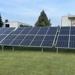Trzy rzędy paneli słonecznych na metalowych nogach, ustawione na zielonym trawniku przed budynkami mieszkalnymi w słoneczny dzień.