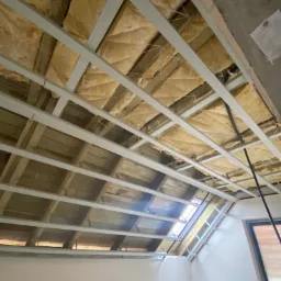 Poddasze w trakcie izolacji, widoczne drewniane belki, wełna mineralna i metalowa konstrukcja stelaża pod przyszłą zabudowę, okno dachowe w tle.