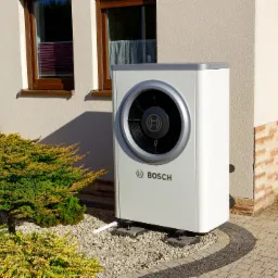 Biała jednostka zewnętrzna z logo Bosch, zamontowana na podłożu z kamyków obok iglastego krzewu i ściany budynku z oknem.