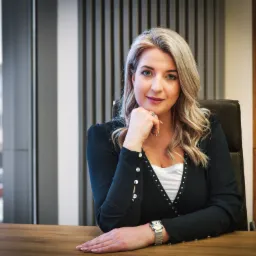 Profesjonalna blondynka w czarnej marynarce z srebrnymi guzikami i zegarkiem na ręce, siedząca za drewnianym biurkiem na tle okna w biurze.