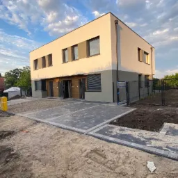 Nowoczesny, dwukondygnacyjny dom z płaskim dachem, wykończony w odcieniach beżu i szarości, z częściowo ukończonym brukowanym podjazdem i ogrodzeniem w tle.