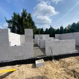 Wznoszenie ścian parterowego domu z białych bloczków, widoczny świeży fundament, poziomica i szpadel na placu budowy w słoneczny dzień.