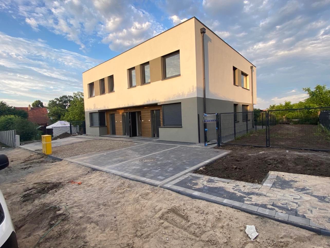 Nowoczesny, dwukondygnacyjny dom z płaskim dachem, wykończony w odcieniach beżu i szarości, z częściowo ukończonym brukowanym podjazdem i ogrodzeniem w tle.