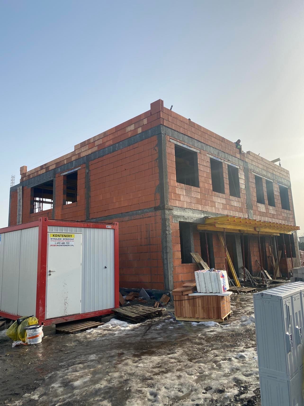 Dwukondygnacyjny budynek w stanie surowym otwartym z czerwonej cegły, z widocznym kontenerem budowlanym i materiałami budowlanymi na placu budowy, w tle lekko zachmurzone niebo.