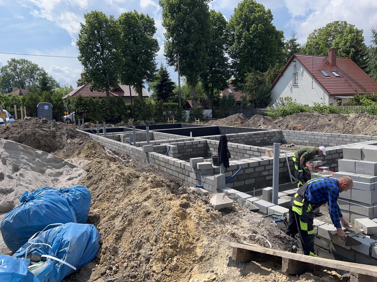 Budowa fundamentów domu: dwóch murarzy w trakcie układania bloczków betonowych, widoczne pionowe rury kanalizacyjne, wykop, hałdy ziemi, worki z materiałami budowlanymi oraz fragmenty zabudowy...