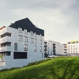 Nowoczesny budynek wielorodzinny z białymi ścianami, czarnymi elementami dachu i balkonami, otoczony zielonym trawnikiem i drzewami, z podziemnym garażem.