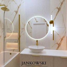 Szymon Jankowski - Elegancka łazienka z marmurowymi płytkami, złotą armaturą i okrągłym lustrem z podświetleniem LED. Minimalistyczny design, nowoczesne oświetlenie.