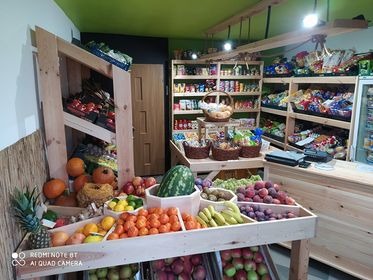 Wnętrze sklepu spożywczego z drewnianymi regałami na owoce i warzywa, widoczne arbuzy, pomarańcze, jabłka, banany i inne produkty, zielony sufit, różne towary na półkach w tle.