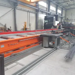 Wnętrze hali produkcyjnej z maszyną do obr&oacute;bki pręt&oacute;w, w tle zaparkowana ciężar&oacute;wka i składowane materiały.