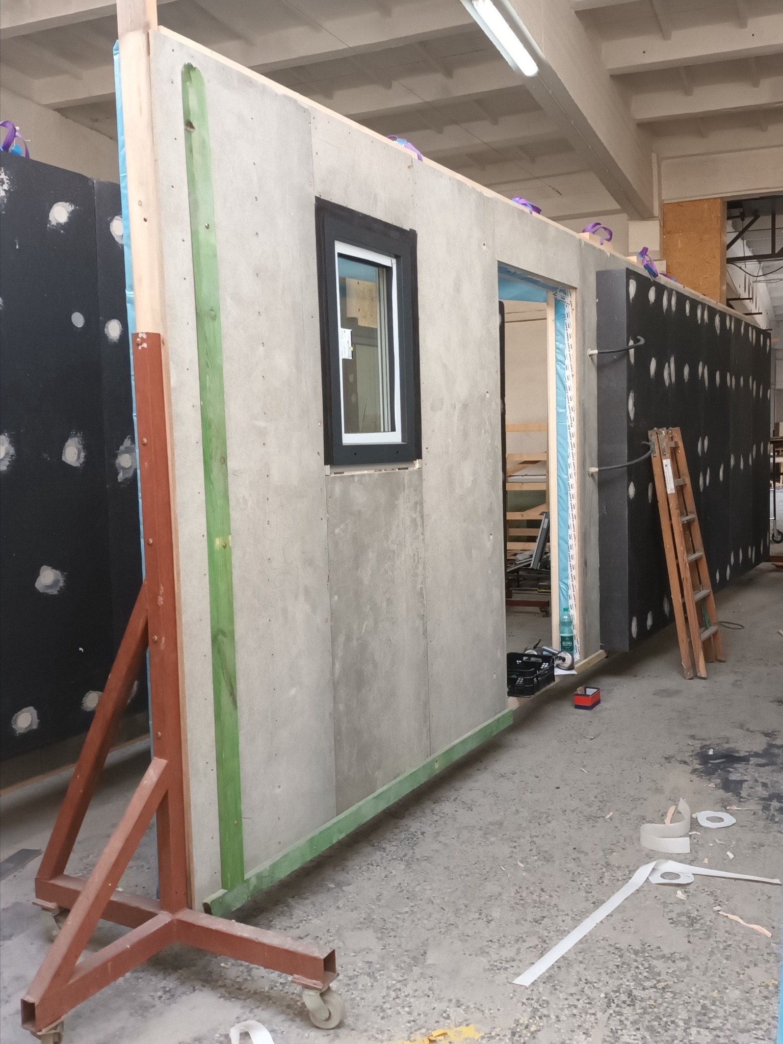 Prefabrykowana ściana modułowa z oknem, oparta na metalowym stojaku na kółkach, widoczne elementy konstrukcyjne i izolacyjne, w tle drabina oparta o ścianę z izolacją.