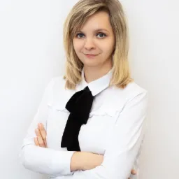 Portret kobiety z blond włosami, ubrana w białą koszulę z czarną kokardą i spódnicę w pepitkę, z założonymi rękami na tle białej ściany.