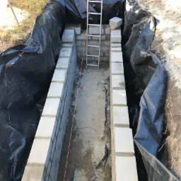 Fundament z bloczków betonowych w wykopie wyłożonym czarną folią, widoczna drabina i naprężony sznurek murarski.