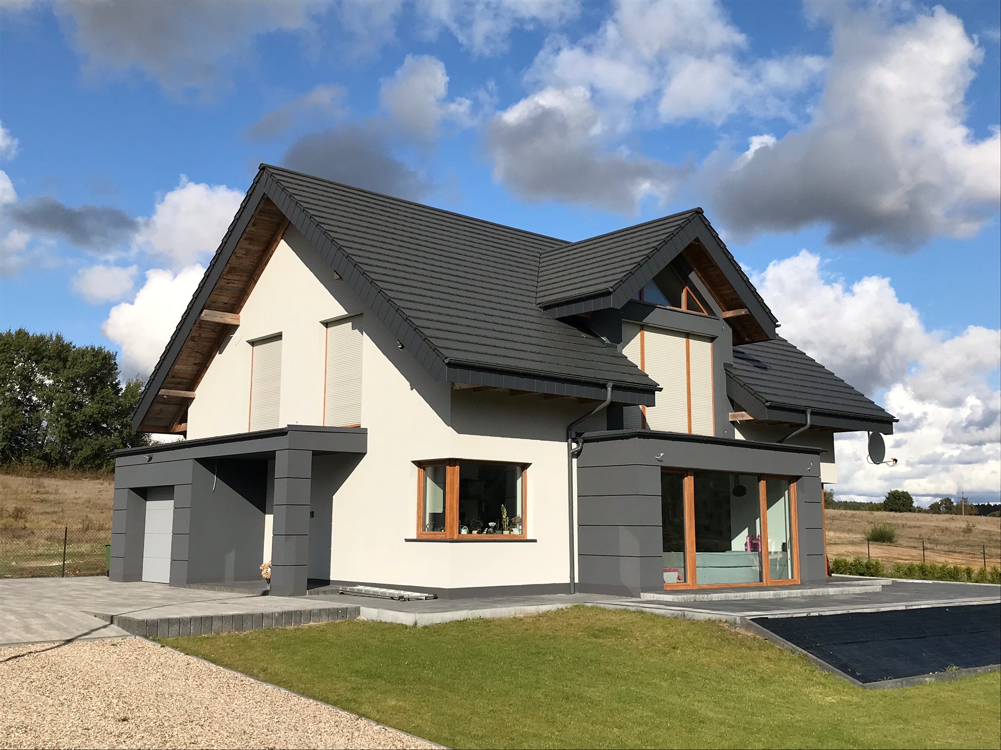 Nowoczesny dom jednorodzinny z grafitowym dachem i jasną elewacją, kontrastujące szare elementy architektoniczne, zadbany trawnik i podjazd z kostki brukowej, błękitne niebo z chmurami w tle.