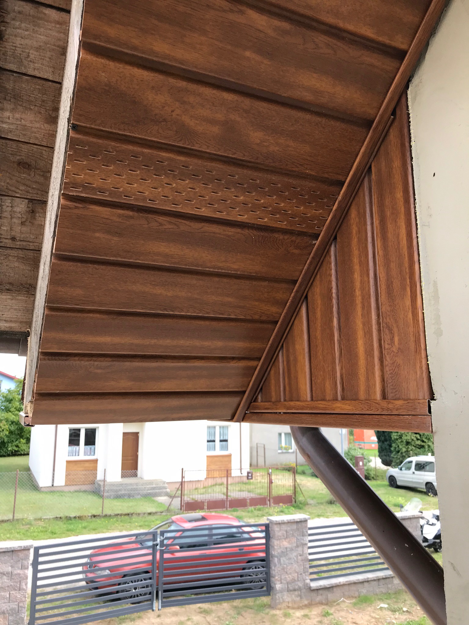 Brązowa podbitka dachowa z imitacją drewna zamontowana na okapie, widok z dołu na tle domu i samochodu.