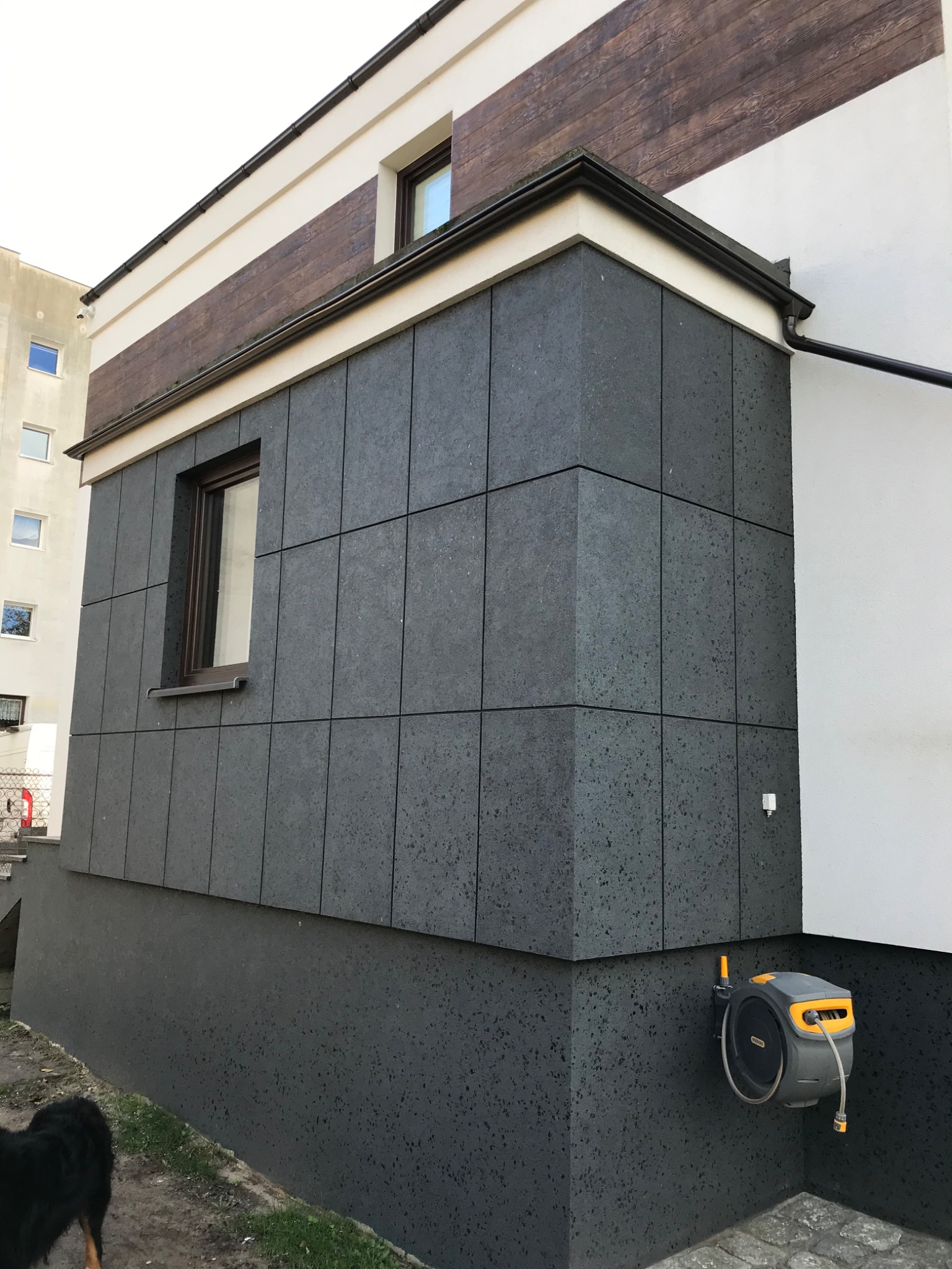 Częściowo wykończona elewacja budynku z szarych, kwadratowych płyt kamiennych, widoczne okno z brązową ramą, zautomatyzowany zwijacz węża ogrodowego zamontowany na ścianie, fragment elewacji...
