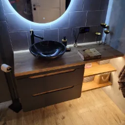 Nowoczesna łazienka z umywalką w kolorze czarnego marmuru, podświetlanym lustrem LED i drewnianym blatem z szufladami, uzupełniona o dekoracyjne świeczniki i zapachowe patyczki.