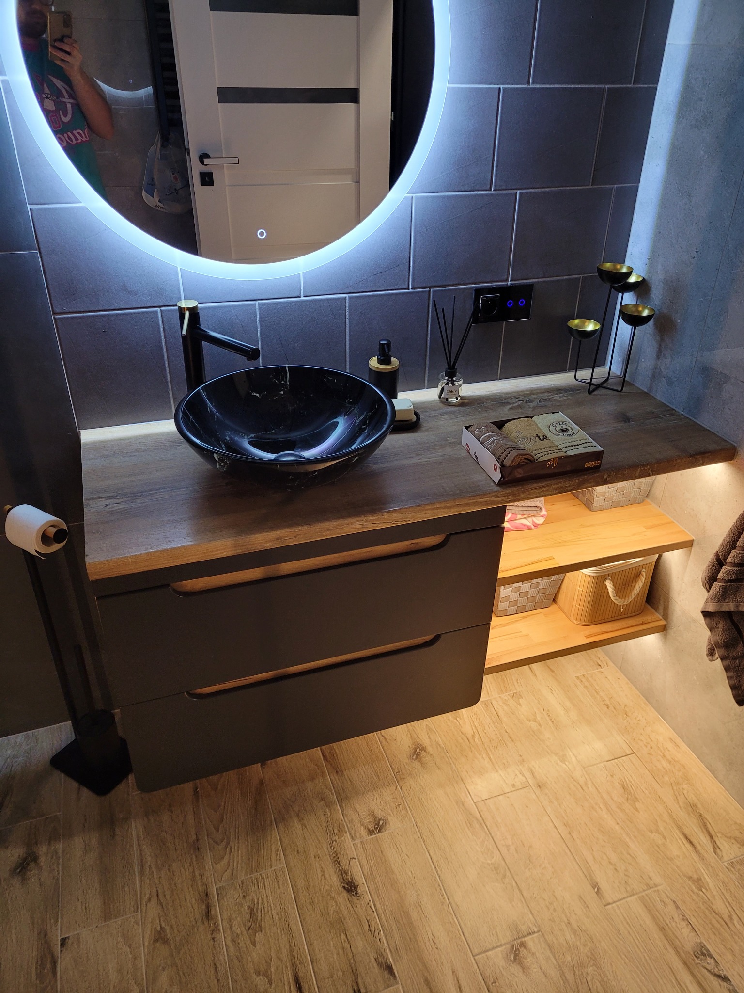Nowoczesna łazienka z umywalką w kolorze czarnego marmuru, podświetlanym lustrem LED i drewnianym blatem z szufladami, uzupełniona o dekoracyjne świeczniki i zapachowe patyczki.