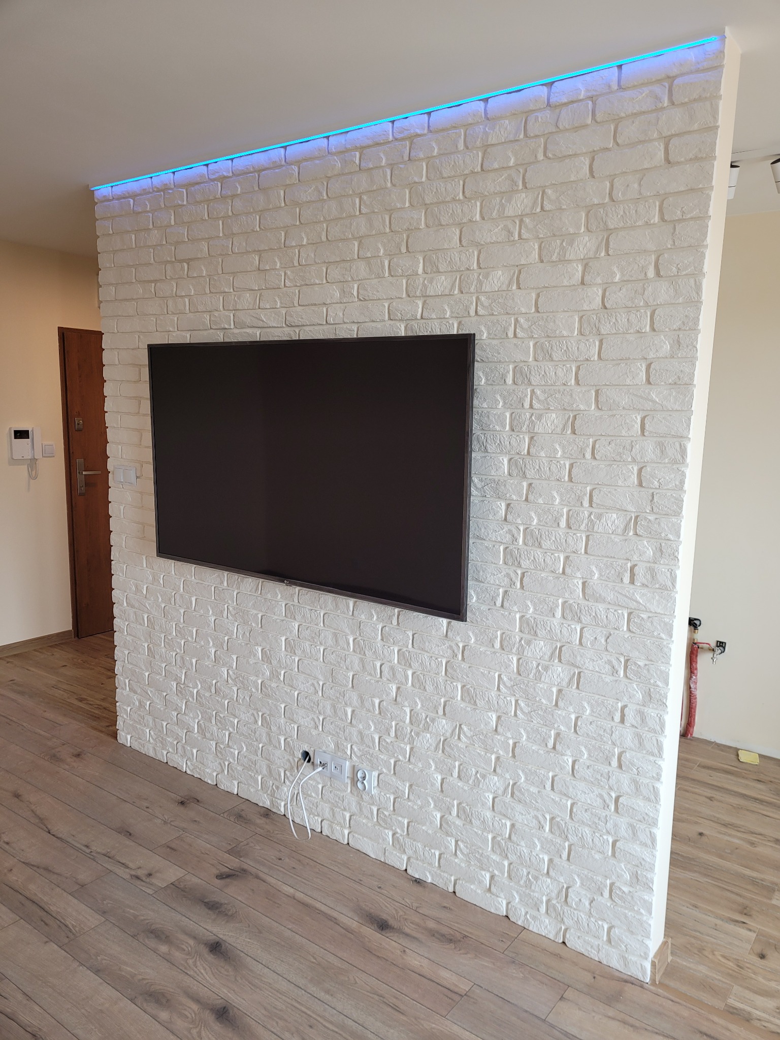 Ściana działowa wykończona białą cegłą dekoracyjną z podświetleniem LED, zamontowany telewizor, drewniana podłoga.