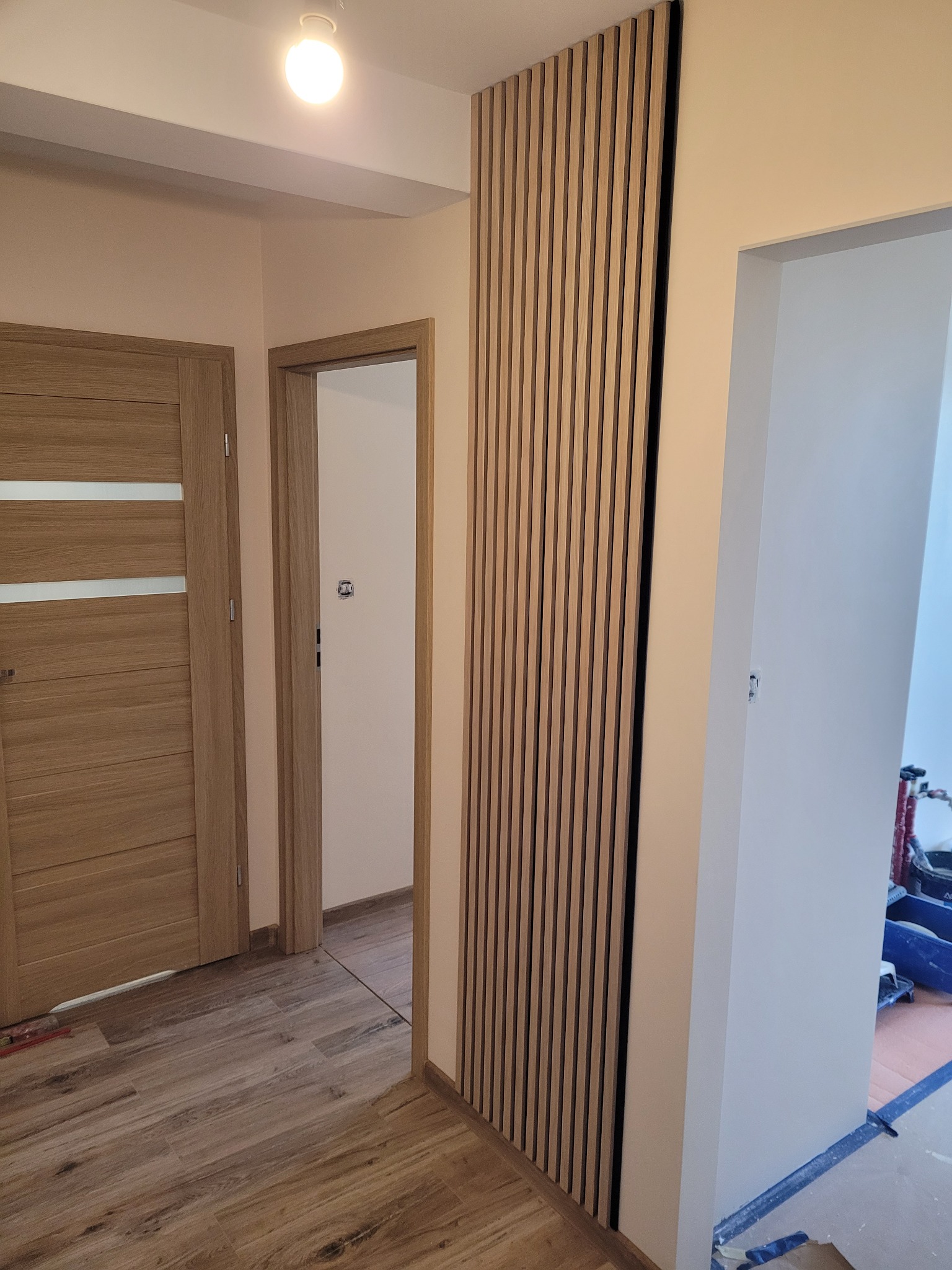 Drewniane lamele ścienne w przedpokoju, widoczne drzwi z poziomymi przeszkleniami, podłoga z paneli imitujących drewno, sufit z oświetleniem punktowym.