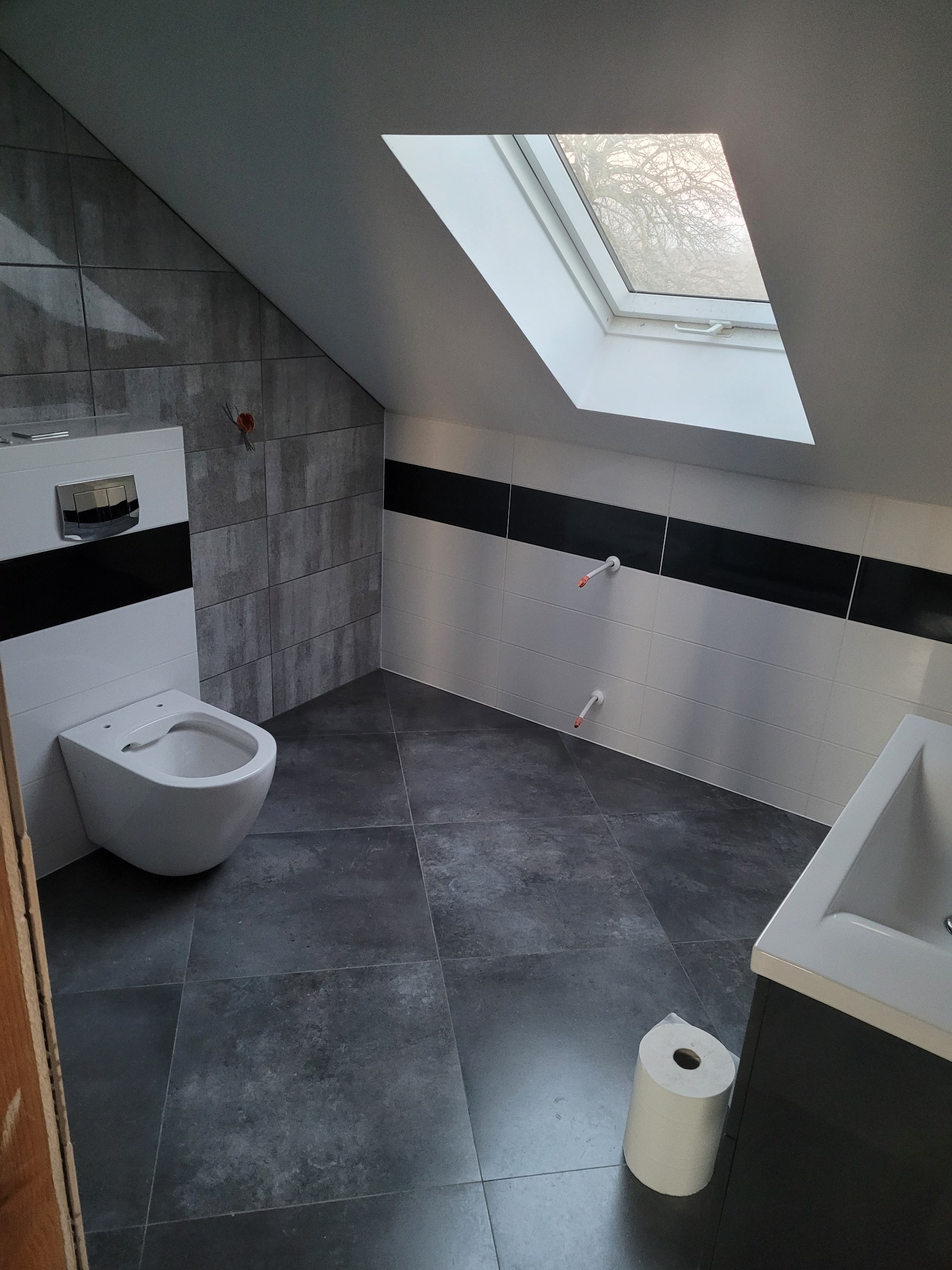 Nowoczesna łazienka na poddaszu z szarymi płytkami podłogowymi i ściennymi, toaleta podwieszana, umywalka z szafką, okno dachowe wpuszczające naturalne światło, widoczne instalacje wodne i rolka...