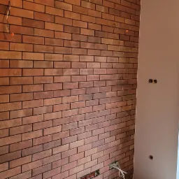 Ściana wyłożona cegiełkami klinkierowymi w odcieniach czerwieni i brązu, widoczne gniazdka elektryczne i wystające kable, okno z widokiem na zieleń na zewnątrz.